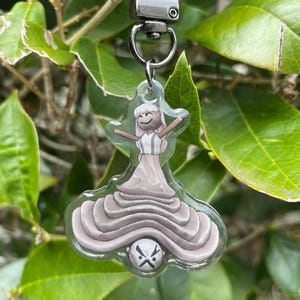 May include: A clear acrylic keychain featuring a cartoon figure with arms outstretched. The figure is wearing a white dress with a light brown skirt. The keychain has a silver-colored metal clasp and is hanging in front of green foliage.