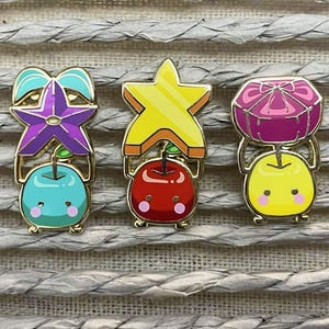 Może przedstawiać: Trzy emaliowane przypinki ze złotymi obwódkami. Każda przypinka ma inny wzór: gwiazdę, pudełko na prezent i gwiazdę. Przypinki są trzymane przez uroczą postać jabłka z różowymi policzkami. Gwiazda jest żółta, pudełko na prezent jest różowe, a gwiazda jest fioletowa.