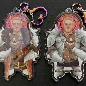 Puede incluir: Dos llaveros acrílicos con un personaje rubio con armadura y abrigo de piel, sosteniendo un pequeño animal marrón. Cada llavero tiene un cierre iridiscente. El fondo presenta un diseño de vidriera.