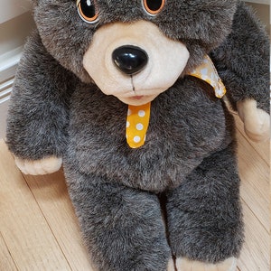 May include: A plush bear toy with dark gray fur, a light beige muzzle, and large orange eyes with black pupils. The bear features a black plastic nose and wears a yellow neck scarf decorated with a white polka dot pattern. The paws are light beige.