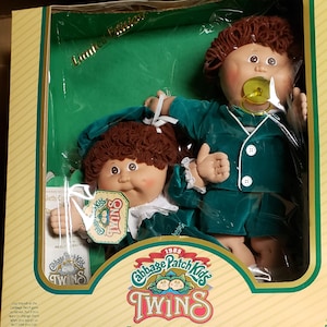 May include: A 1985 Limited Edition Cabbage Patch Kids Twins doll set in original packaging. The two dolls feature brown yarn hair, round faces, and matching dark green velvet outfits. One doll has a yellow pacifier, and the box displays the text Cabbage Patch Kids Twins and Twins are twice as nice.