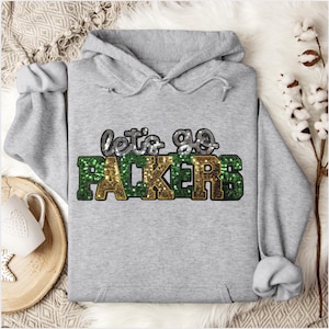 May include: A gray hooded sweatshirt with the text "let's go PACKERS" in sequined lettering. The word "PACKERS" is in green and gold sequins. The sweatshirt has a drawstring hood and long sleeves.