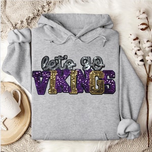 May include: A gray hooded sweatshirt with the text "let's go VIKINGS" in silver, purple, and gold sequins. The sweatshirt has a drawstring hood and long sleeves. The background includes a white fluffy surface and a wooden tray with a mug.