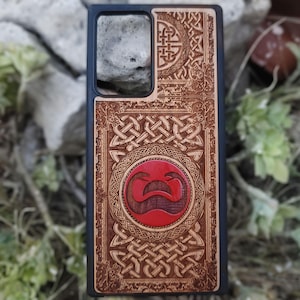 Puede incluir: Funda de teléfono negra con intrincados diseños de nudos celtas grabados en madera. La funda presenta un emblema rojo central y patrones detallados, combinando arte y protección.