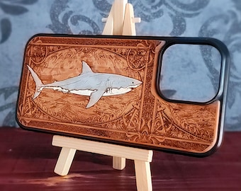 May include: A rectangular phone case with a black frame and a light brown wood-toned back panel. The back features a laser-engraved illustration of a shark swimming within an oval border, surrounded by intricate decorative patterns and anchor motifs.