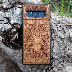 May include: A wooden phone case featuring a laser-engraved design of a spider in a web. The case has a black border and a camera cutout. The text "Goliath Bird Eater" is engraved at the bottom of the case.