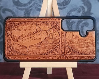 May include: A rectangular phone case with a black frame and a light brown wood-toned back panel. The back features an intricate laser-engraved design of a marlin fish swimming in the ocean, accompanied by sailing ships and anchor motifs.
