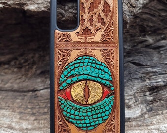 May include: A wooden phone case with a carved design of a green eye with red and yellow accents. The eye is surrounded by a decorative frame.