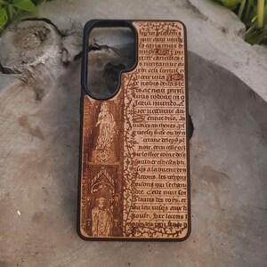 May include: A wooden phone case with a black border. The case features an intricate design of engraved text and illustrations, including figures in a gothic style. The wood has a warm, natural tone, and the design appears to be inspired by medieval manuscripts.