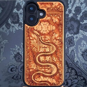 Może przedstawiać: Drewniane etui na telefon z szczegółowym, grawerowanym wzorem. Etui przedstawia węża wijącego się przez kwiatowy wzór, z ciemną obwódką. Zaprojektowane dla smartfona z podwójnym obiektywem aparatu.