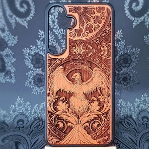 Könnte beinhalten: Eine Handyhülle aus Holz mit einem detaillierten, geschnitzten Design. Die Hülle zeigt ein zentrales Bild eines Vogels mit ausgebreiteten Flügeln, umgeben von Zierornamenten und einem Berg am Boden. Schwarzer Rand.