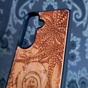 May include: A black smartphone case featuring a light brown wood inlay on the back. The wood surface is laser-engraved with a detailed illustration of an octopus with visible suction cups and a portion of a vintage diving helmet at the bottom.