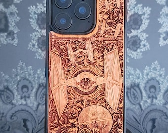 May include: A wooden phone case with a detailed laser-etched design. The design features a TIE fighter, the Death Star, and other elements from a popular science fiction franchise. The case has a black border and a natural wood colour.