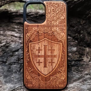 May include: A wooden phone case with a black border and a carved design of a shield with four crosses. The shield is surrounded by a decorative floral pattern.