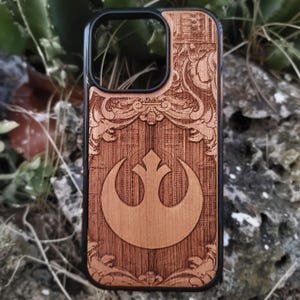 May include: A wooden phone case with a black border. The case features a carved design with a prominent emblem and decorative scrollwork. The emblem is a stylized crescent and cross design. The wood has a warm, natural tone.
