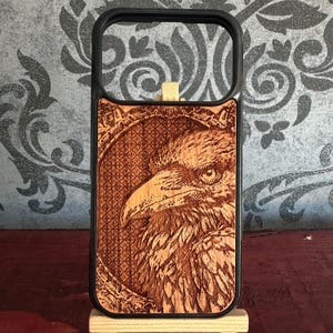 May include: A rectangular phone case featuring a black plastic frame and a light brown wooden back panel. The wood is laser-engraved with a detailed portrait of a raven inside an ornate, patterned oval frame with intricate scrollwork.