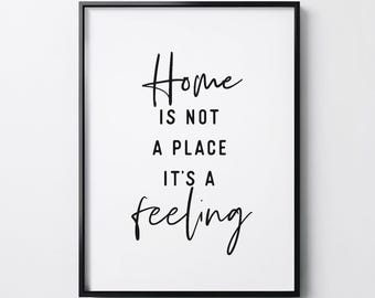 May include: A rectangular wall art print in a thin black frame featuring a white background with black text that reads: Home IS NOT A PLACE IT'S A feeling. The word Home and the word feeling are written in a cursive script, while the rest is sans-serif.