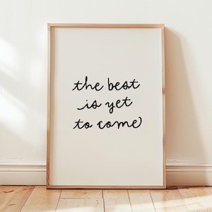 May include: A framed print with the phrase "the best is yet to come" in black script. The print has a cream-colored background and a light brown wooden frame. The artwork is leaning against a white wall on a wooden floor.