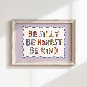Op de afbeelding: Een ingelijste kunstdruk met een crèmekleurige, golvende rechthoek op een lichtpaarse achtergrond. De tekst luidt BE SILLY, BE HONEST, BE KIND in veelkleurige, handgetekende letters. De lijst is gemaakt van wit hout met een verweerde look.