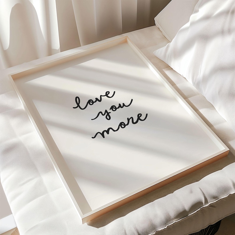 May include: A rectangular framed wall art piece featuring a white background with the black cursive text "love you more" centered on the print. The frame is light-colored wood, and the artwork is displayed in a clean, simple orientation.