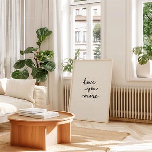 May include: A rectangular framed wall art piece featuring the black handwritten text "love you more" centered on a white background. The frame is a light-toned wood, and the artwork is positioned upright on a floor next to a wooden coffee table.