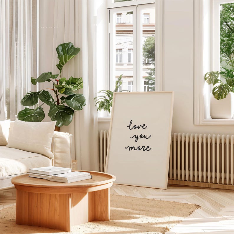May include: A rectangular framed wall art piece featuring the black handwritten text "love you more" centered on a white background. The frame is a light-toned wood, and the artwork is positioned upright on a floor next to a wooden coffee table.