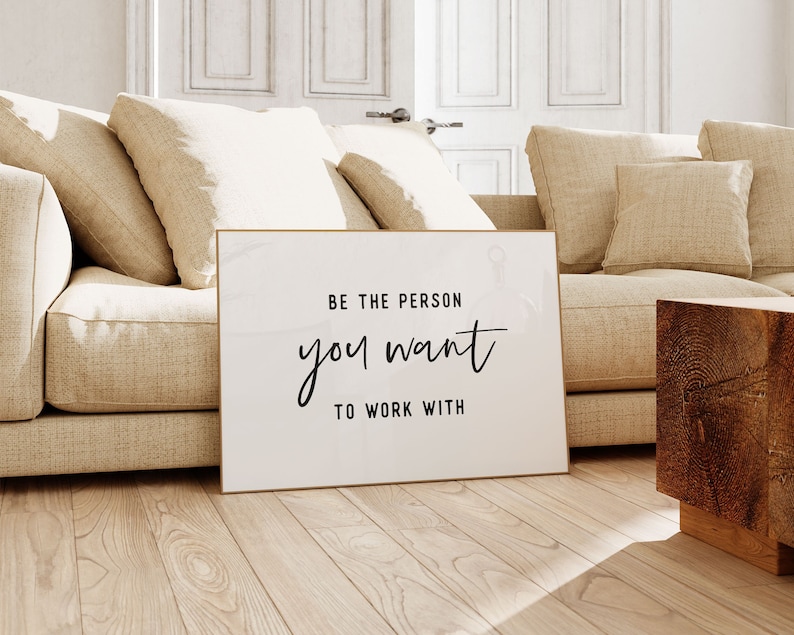 Pu&ograve; includere: Un poster bianco incorniciato con il testo "Be the person you want to work with" in corsivo nero. Il poster &egrave; appoggiato a un divano beige in un soggiorno.