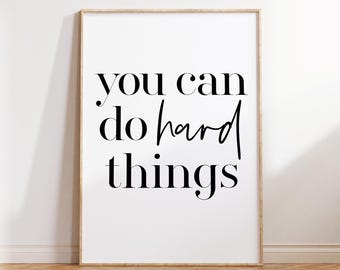 Puede incluir: Impresión enmarcada con la frase "you can do hard things" en escritura negra sobre fondo blanco. El marco es de madera clara, y la impresión se muestra contra una pared blanca y suelo de madera.