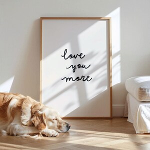 May include: A rectangular wall art print in a light wood frame featuring the black handwritten text "love you more" on a white background. The print is positioned on a wooden floor next to a sleeping golden-colored dog and a white upholstered piece of furniture.
