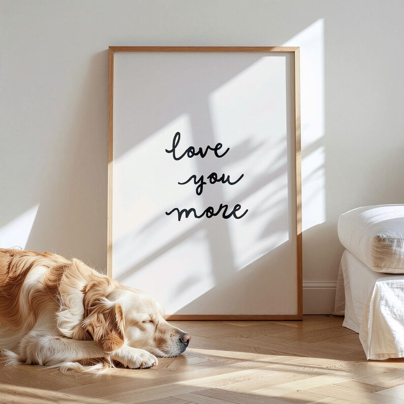 May include: A rectangular wall art print in a light wood frame featuring the black handwritten text "love you more" on a white background. The print is positioned on a wooden floor next to a sleeping golden-colored dog and a white upholstered piece of furniture.