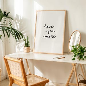 May include: A rectangular framed wall art piece featuring the black handwritten text "love you more" centered on a white background. The frame is made of light-colored wood and is positioned on a white desk surface next to a small potted cactus.