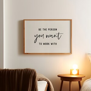 Pu&ograve; includere: Una citazione motivazionale incorniciata su una parete bianca recita "Sii la persona con cui vuoi lavorare". La citazione &egrave; in corsivo nero. La cornice &egrave; in legno chiaro. Una coperta marrone &egrave; su un letto in primo piano. Un tavolino con una lampada si trova nell'angolo destro dell'immagine.