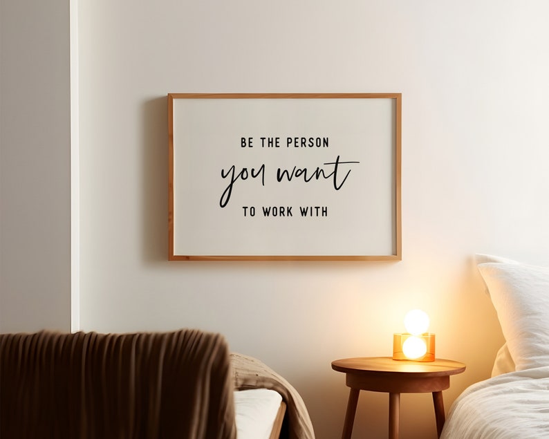 Pu&ograve; includere: Una citazione motivazionale incorniciata su una parete bianca recita "Sii la persona con cui vuoi lavorare". La citazione &egrave; in corsivo nero. La cornice &egrave; in legno chiaro. Una coperta marrone &egrave; su un letto in primo piano. Un tavolino con una lampada si trova nell'angolo destro dell'immagine.