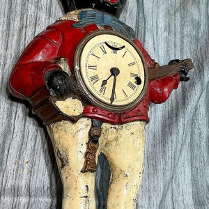 Könnte beinhalten: Eine Vintage-Gusseisen-Uhrfigur, die eine Person mit Zylinder, roter Jacke und cremefarbener Hose darstellt. Die Figur hält ein rundes Zifferblatt mit römischen Ziffern auf der Brust und steht auf einem rechteckigen Sockel mit abgenutzter Oberfläche.