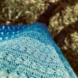 Könnte beinhalten: Nahaufnahme eines blau-türkisfarbenen gehäkelten Schals. Der Schal hat ein strukturiertes, offenes Webmuster. Die Farbe geht von einem tiefen Blau oben zu einem helleren Türkis unten über. Der Hintergrund ist verschwommen und zeigt grünes Gras.