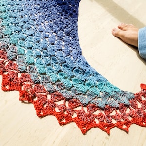 Peut inclure: Un châle fait à la main, crocheté dans un dégradé de couleurs, passant du bleu profond au turquoise et enfin au rouge vif. Le bord du châle présente un motif festonné. L'article est posé sur une surface en bois de couleur claire.
