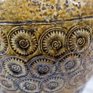 May include: A brown ceramic jar with a textured surface and a repeating pattern of stylized flowers. The jar has a wide mouth and a rounded body.