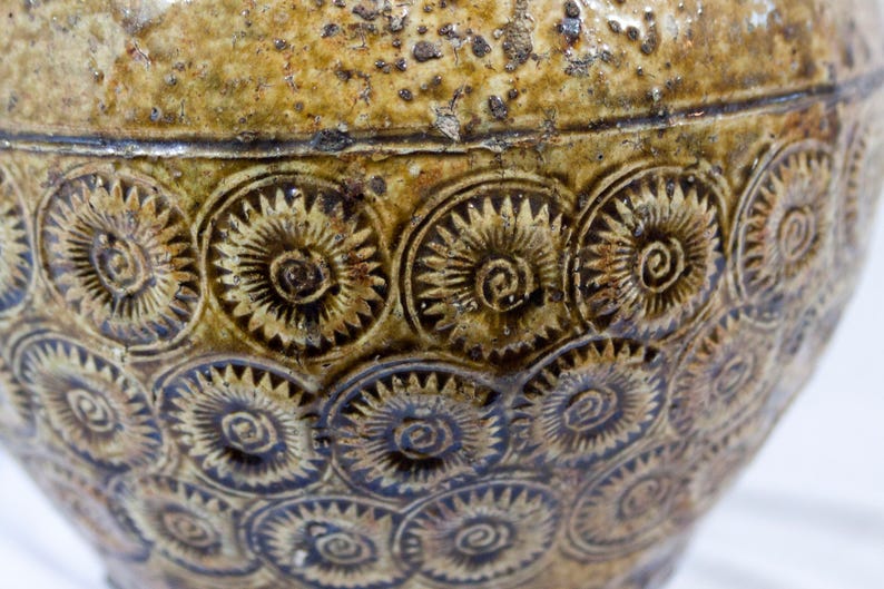 May include: A brown ceramic jar with a textured surface and a repeating pattern of stylized flowers. The jar has a wide mouth and a rounded body.