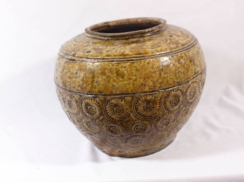 May include: A large, brown ceramic jar with a textured surface and a pattern of raised circles. The jar has a wide mouth and a rounded body.