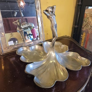 May include: A silver-toned Art Nouveau centerpiece dish. The dish is shaped like a stylized flower with a slender female figure at the center. The figure has her arms raised above her head. The dish is on a dark brown surface.