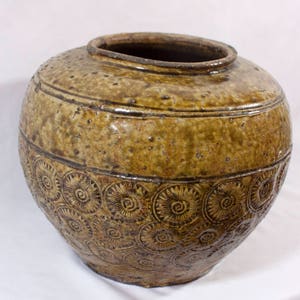 May include: A large, brown ceramic jar with a decorative pattern of swirling circles. The jar has a wide mouth and a rounded body.