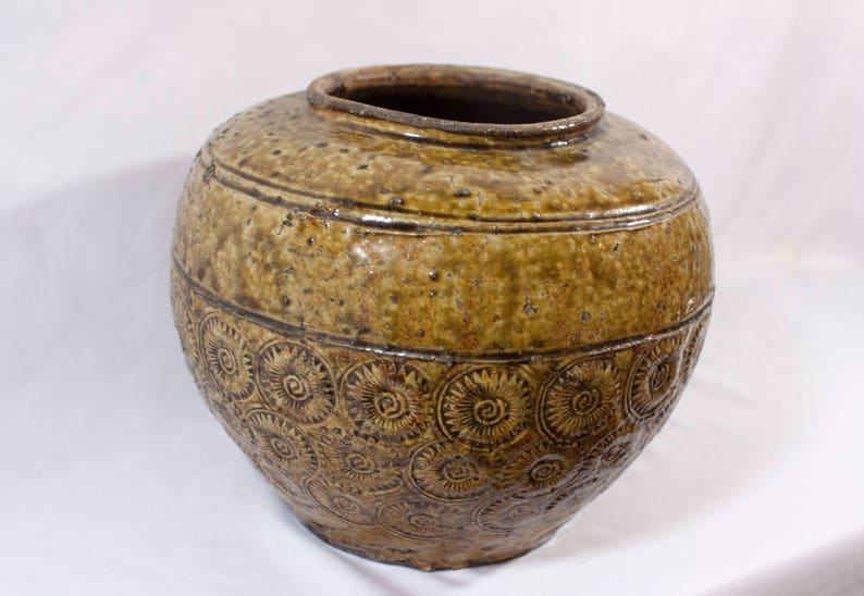 May include: A large, brown ceramic jar with a decorative pattern of swirling circles. The jar has a wide mouth and a rounded body.