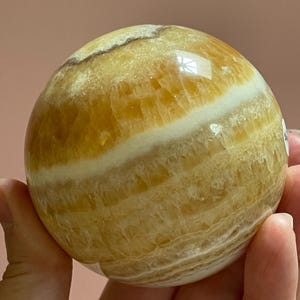 May include: A polished, spherical gemstone with bands of yellow and white. The orb is held in a hand, showcasing its smooth, reflective surface. The stone's natural patterns and colors are clearly visible.