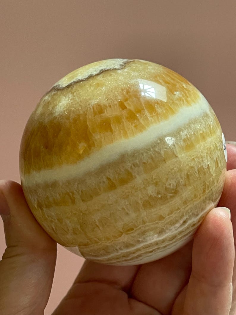 May include: A polished, spherical gemstone with bands of yellow and white. The orb is held in a hand, showcasing its smooth, reflective surface. The stone's natural patterns and colors are clearly visible.