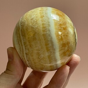 May include: A polished, spherical gemstone with a warm, golden-yellow hue, accented by bands of creamy white. The orb is held in a hand, showcasing its smooth, reflective surface. The stone's natural patterns create a unique, eye-catching design.