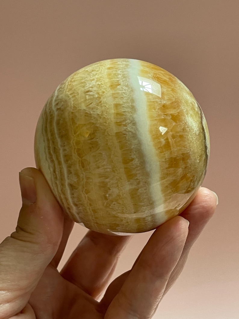 May include: A polished, spherical gemstone with a warm, golden-yellow hue, accented by bands of creamy white. The orb is held in a hand, showcasing its smooth, reflective surface. The stone's natural patterns create a unique, eye-catching design.