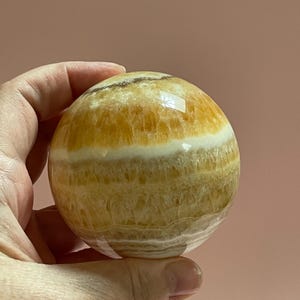 May include: A polished, spherical decorative stone with bands of yellow, white, and tan hues. The orb is held in a hand, showcasing its smooth, reflective surface. The stone's natural patterns and colors create a unique, eye-catching piece.