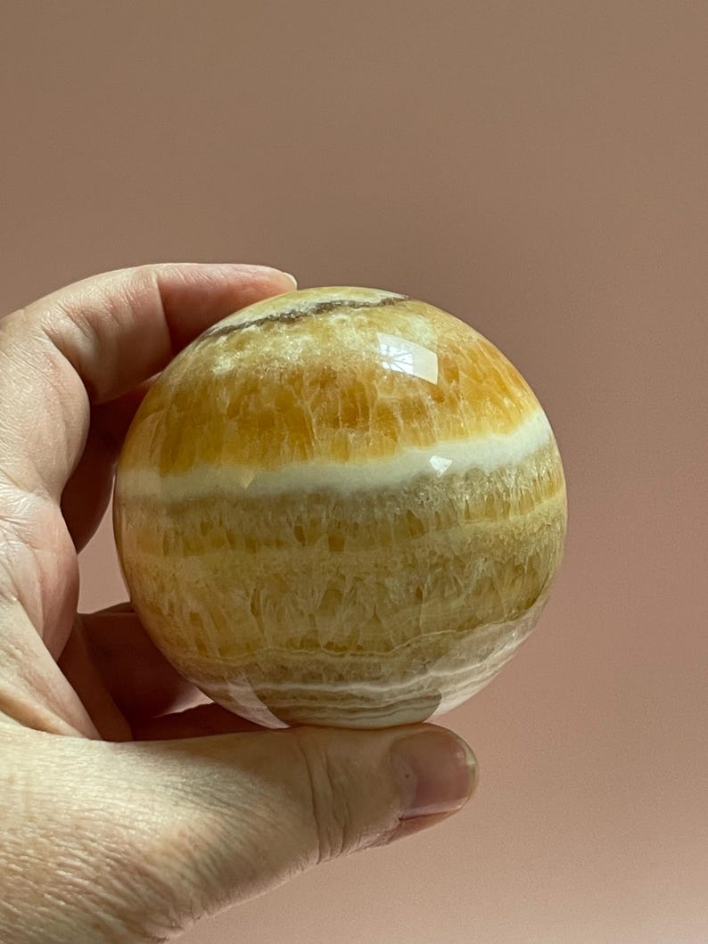 May include: A polished, spherical decorative stone with bands of yellow, white, and tan hues. The orb is held in a hand, showcasing its smooth, reflective surface. The stone's natural patterns and colors create a unique, eye-catching piece.