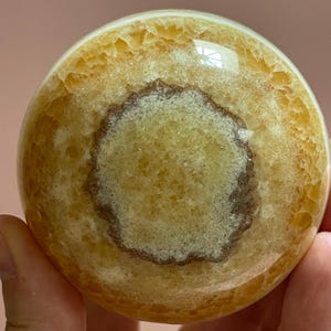 May include: A polished, spherical gemstone with a warm, yellow-orange hue. The orb features a central, circular pattern in a lighter shade, surrounded by a darker, textured band. The surface has a glossy finish, reflecting light.
