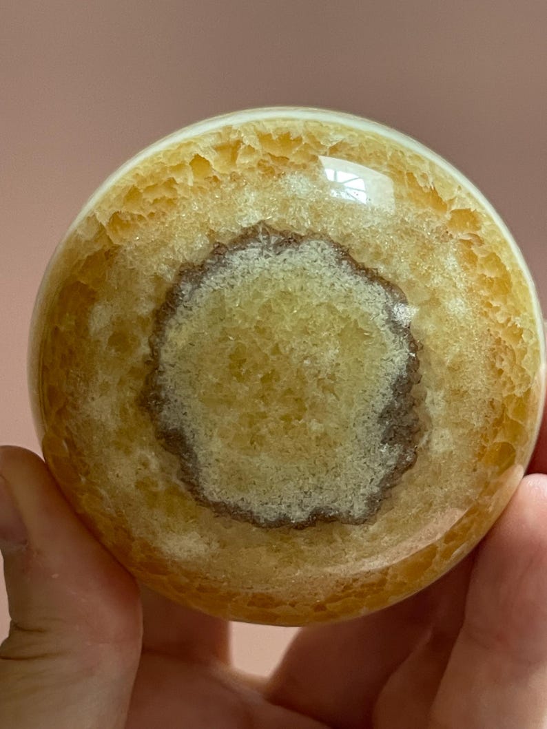 May include: A polished, spherical gemstone with a warm, yellow-orange hue. The orb features a central, circular pattern in a lighter shade, surrounded by a darker, textured band. The surface has a glossy finish, reflecting light.